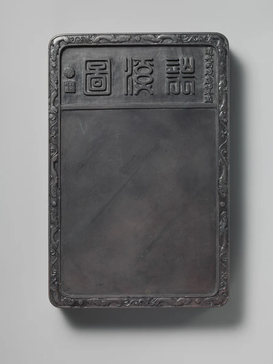 A dark slate inkstone, plain and worn, with seal-script characters cut across its head.